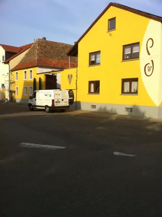 Gelb gestrichenes Wohnhaus mit großem Wandlogo „Schmitt Wein & Sekt“ und sichtbarem Firmenfahrzeug.