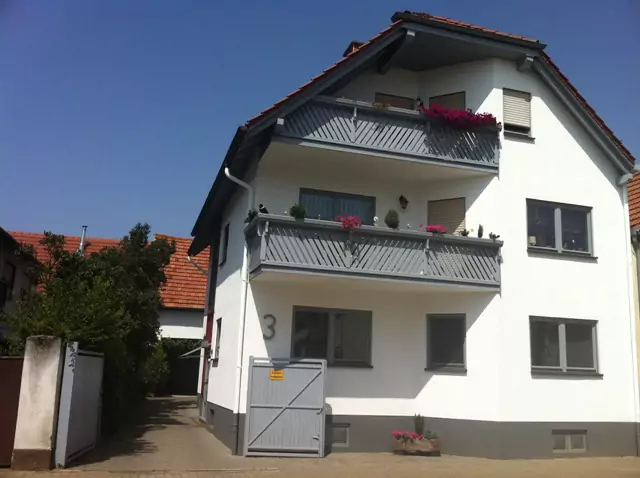 Dreistöckiges Wohnhaus mit grauen Holzbalkonen, Blumenschmuck und frisch gestrichener Fassade.