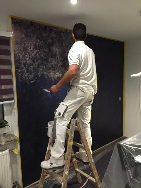 Handwerker auf Leiter gestaltet eine dunkle Wand mit kreativer Spachteltechnik.